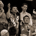 Group of men on a stage at a rally one man speaks into a microphone while others cheer and raise hands
